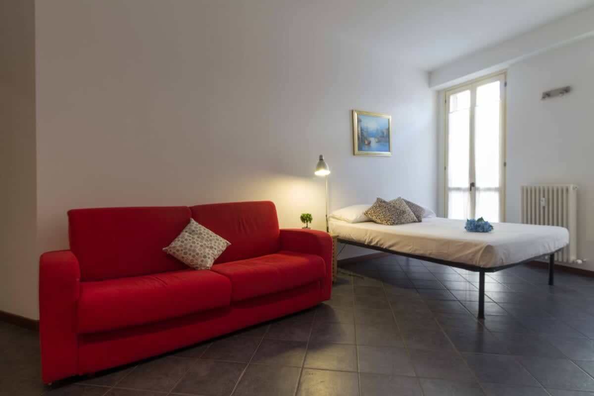 A room with a red sofa, a bed, a small table, a lamp, and a window.