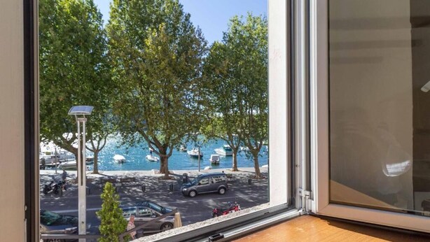A view from a window with a wooden floor, looking out to a parking area with trees and a body of water. - Lungolago Holiday Home - Affitti Brevi Italia (Lecco)