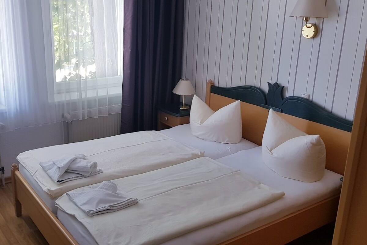 Une chambre d’hôtel avec deux lits, une table de chevet avec une lampe, une chaise et une fenêtre avec des rideaux légers.
