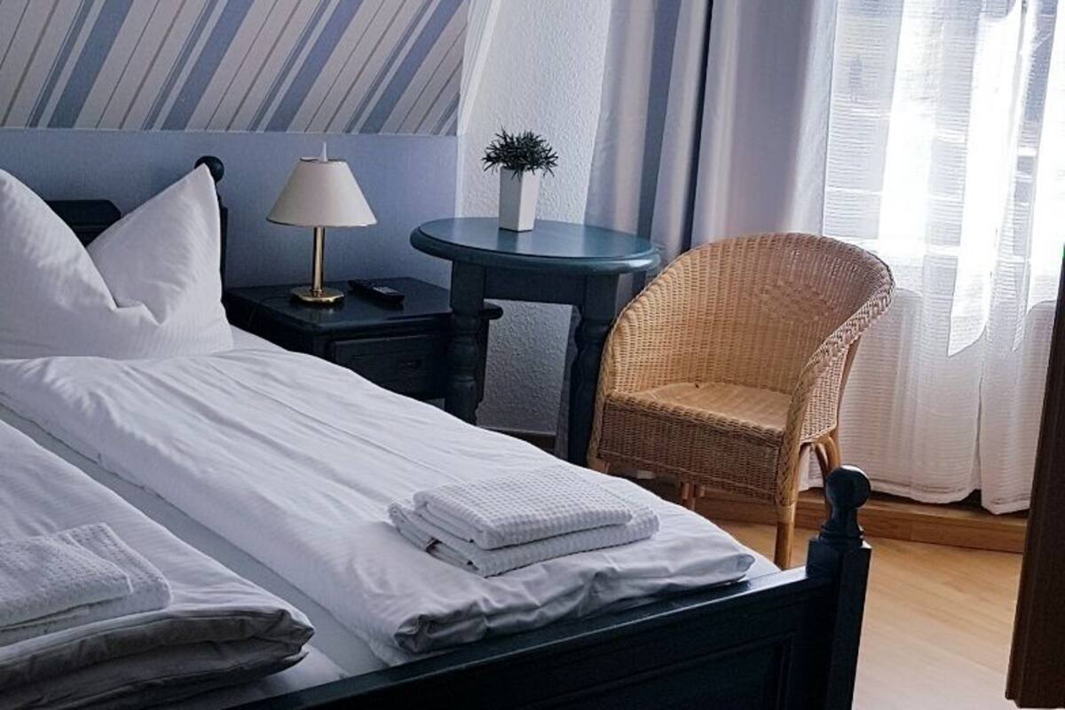 Un lit bien fait, avec du linge de lit blanc, une petite table ronde avec une lampe, une chaise en osier et un mur à rayures.