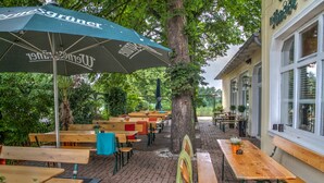Een terras met houten tafels en bankjes, omgeven door bomen en een gebouw met grote ramen.