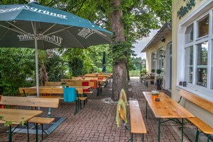 Um café ao ar livre com mesas e bancos de madeira, rodeado por árvores e um edifício com grandes janelas.