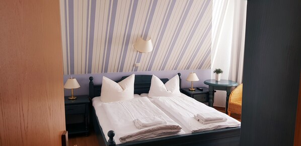 A neatly made bed with white linens and pillows, two bedside tables with lamps, a small round table with a lamp, and a striped wallpaper background. - Restaurant und Pension Zur Nassen Ecke (Wittenbeck)