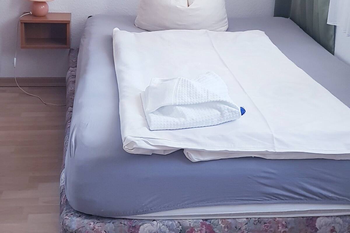 Un lit bien fait, avec une housse de matelas à motifs floraux, une lampe de chevet et une œuvre d’art florale encadrée accrochée au mur.