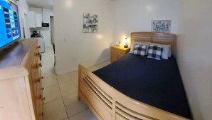 A bedroom with a wooden bed, a nightstand with a lamp, a small kitchen area, and a television on a stand. - #20 Marjory’s in-law suite (Tampa)