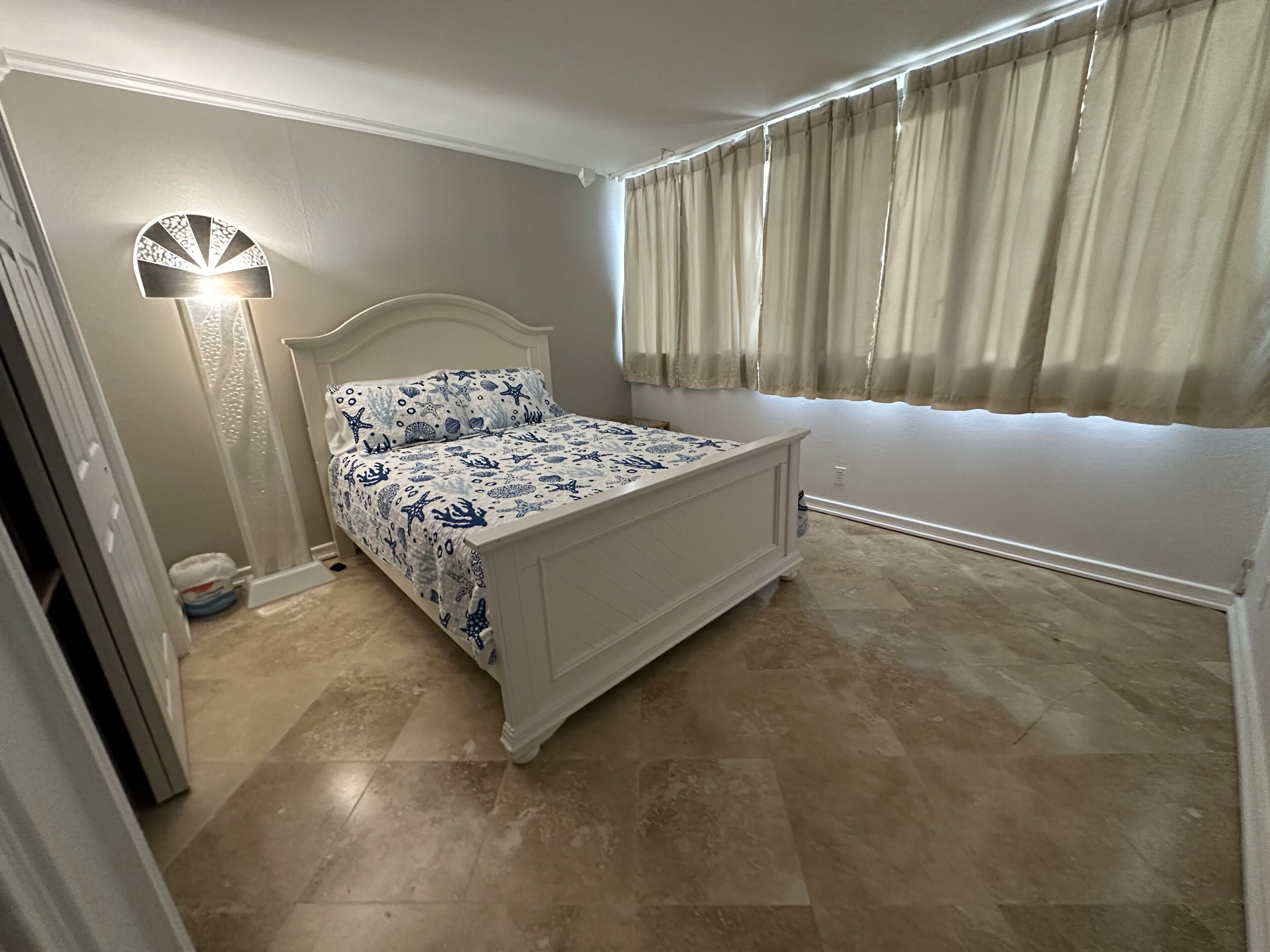 Quarto com cama, mesa de cabeceira, abajur, janela com cortinas e porta.
