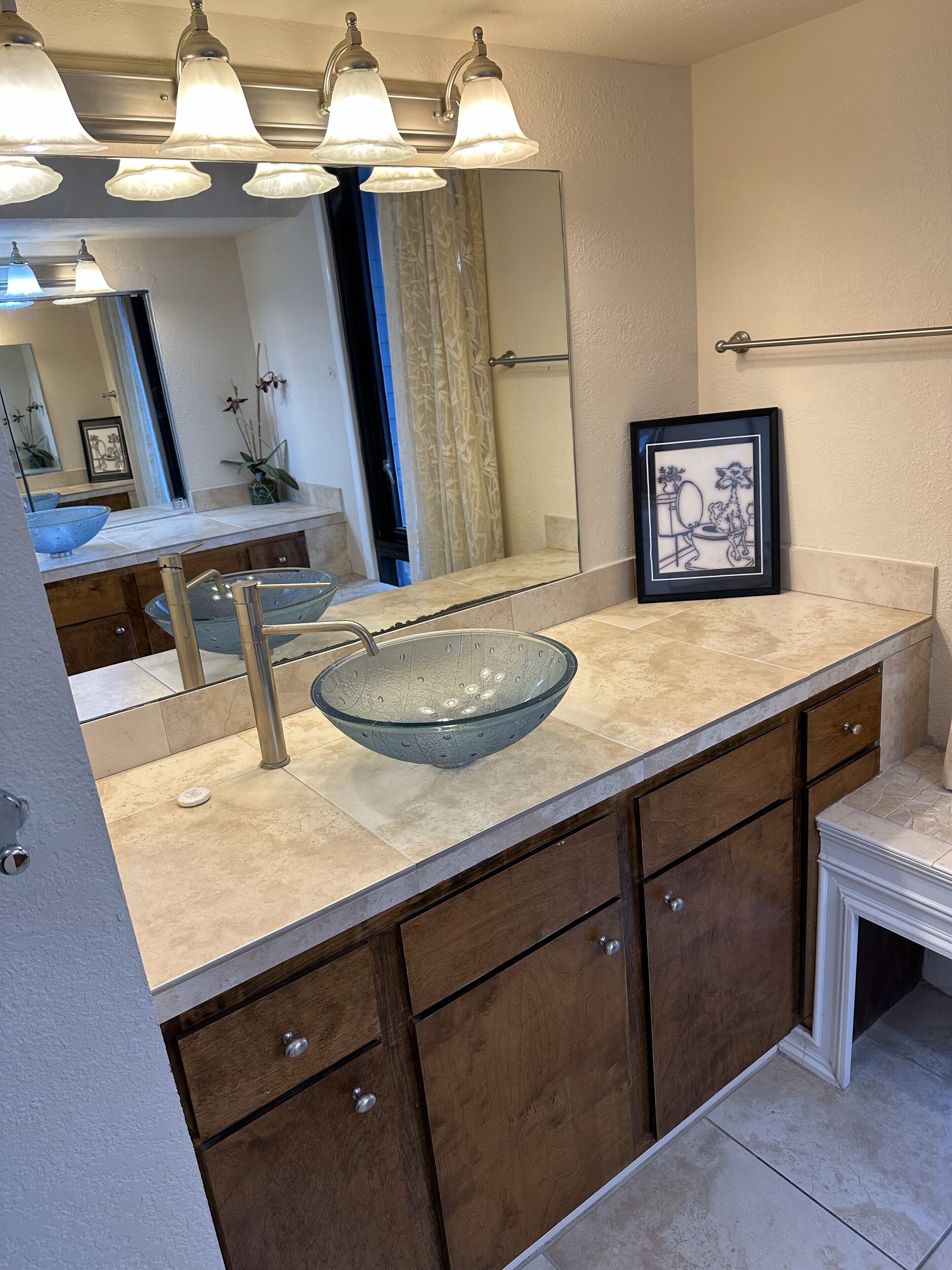 Un baño moderno con un espejo grande, un lavabo de vidrio, armarios de madera y un cuadro en la pared.