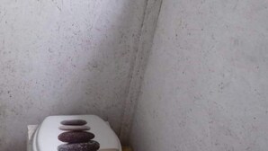 Un baño pequeño y funcional con inodoro blanco, un estante pequeño encima y un paquete de papel higiénico en el suelo.