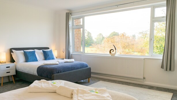 Ein Schlafzimmer mit einem großen Bett, einem Nachttisch, einer Kommode, einem Fenster mit Blick auf Bäume und einer Heizung.