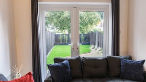 Ein Wohnzimmer mit grauem Sofa, roten und blauen Kissen und einer Glastür, die zu einem Hinterhof mit Holzzaun und viel Grün führt.