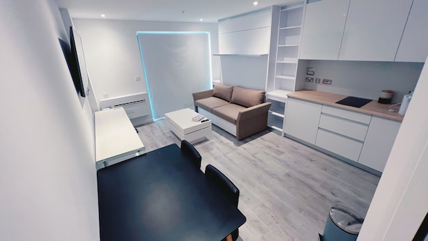 A modern kitchen with white cabinets, a dining table with chairs, a sofa, and a small trash bin. - Modern Studio Apartment Gibraltar (Gibraltar)