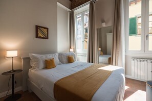 A neatly made bed with white linens and a brown throw, a bedside lamp, and a window with curtains. - Atelier delle Grazie (Florence)