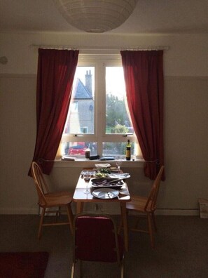 Uma área de refeições com uma mesa posta para dois, duas cadeiras, uma janela com cortinas e vista para uma casa lá fora.