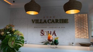 Ett receptionsområde med en skylt som visar "Villa Caribe" och "Soha Beauty Club". På disken står två drinkar och till vänster finns en stor växt.