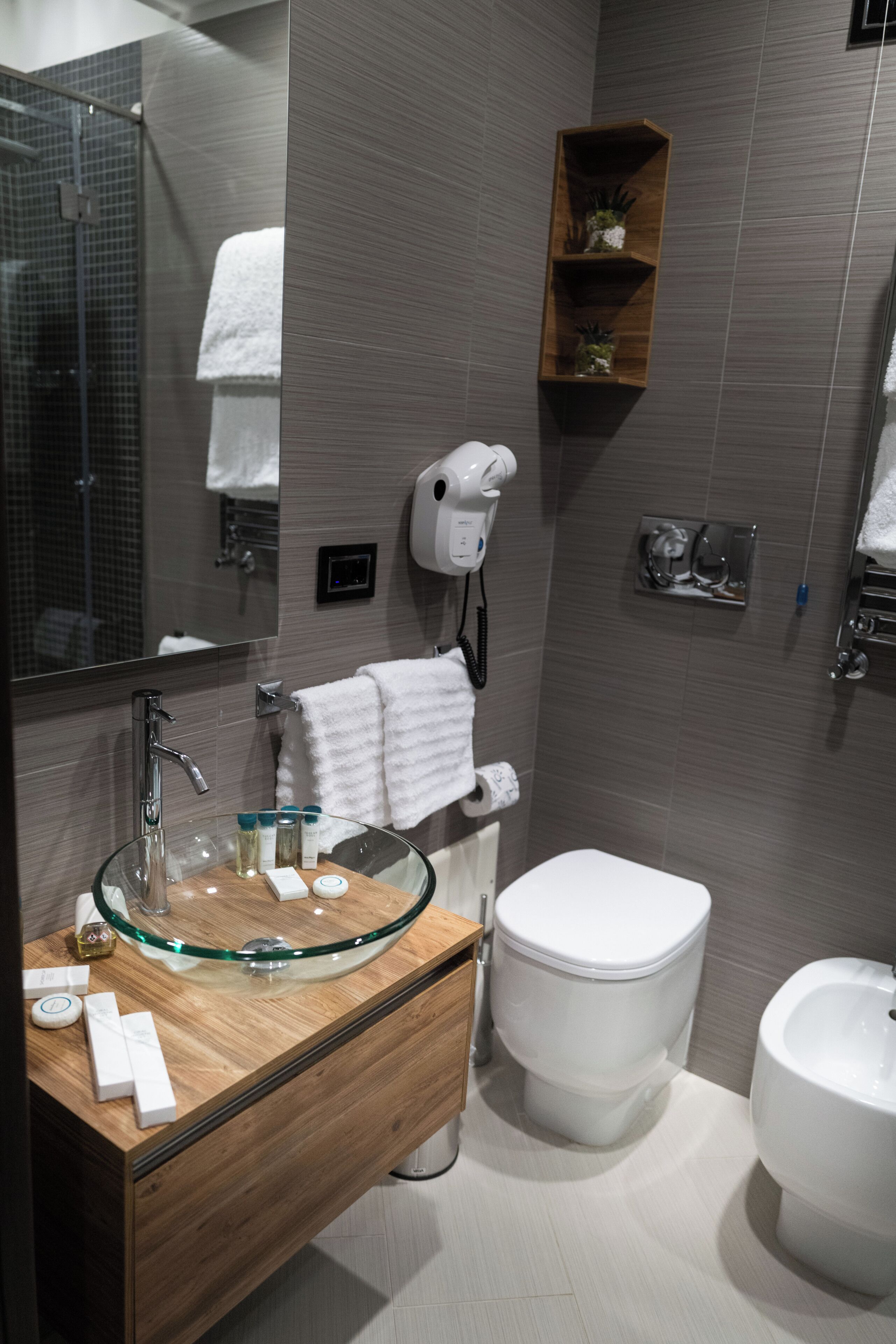 Ein modernes Badezimmer mit Waschbecken aus Glas, Holzwaschtisch, weißer Toilette, Bidet und Handtuchhalter.