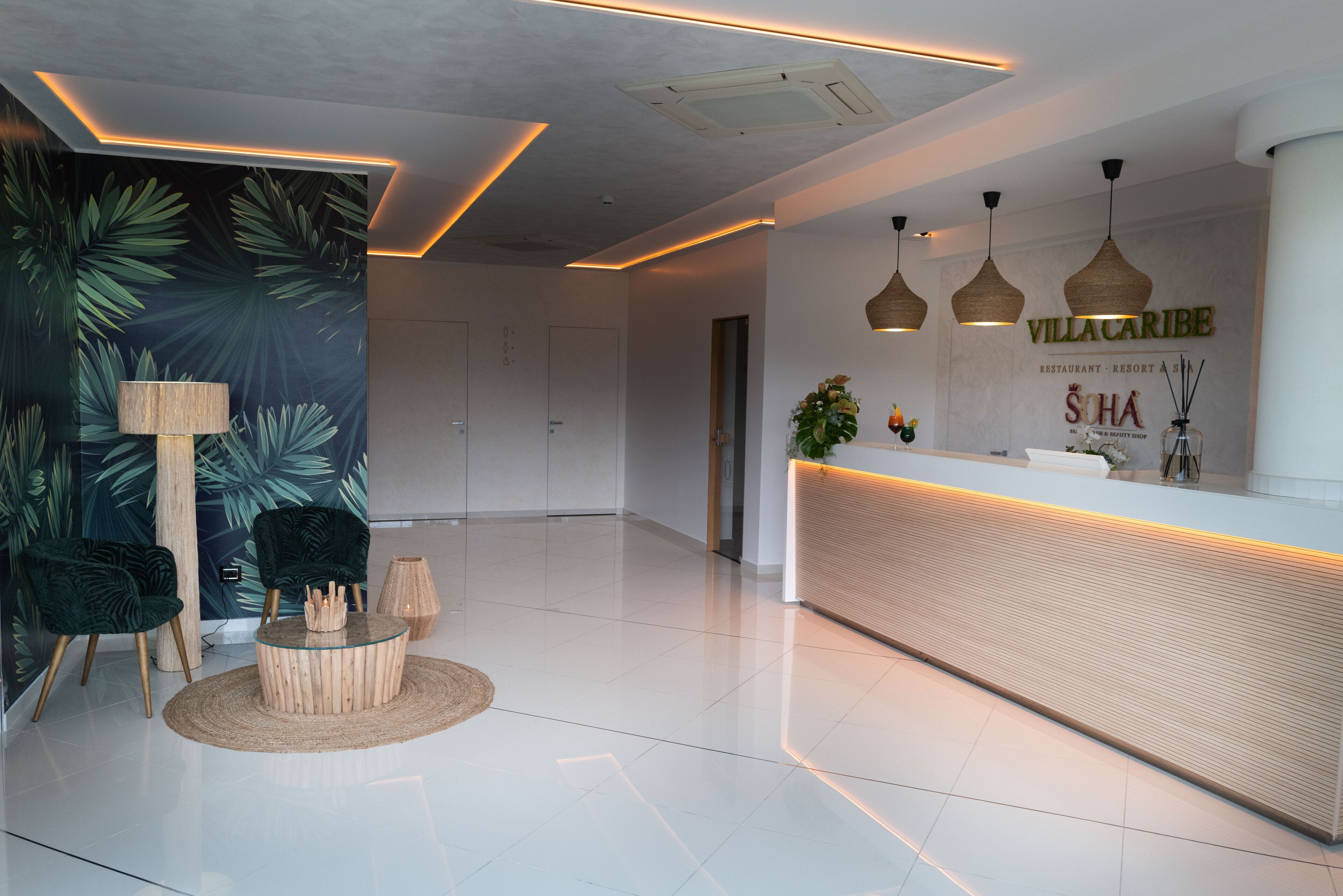 Un área de recepción moderna con un mostrador de madera, dos lámparas colgantes y una pared con estampado de hojas tropicales.