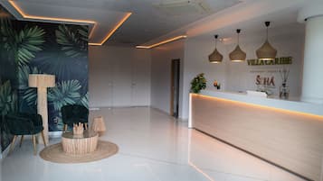 Un área de recepción moderna con un mostrador de madera, dos lámparas colgantes y una pared con estampado de hojas tropicales.