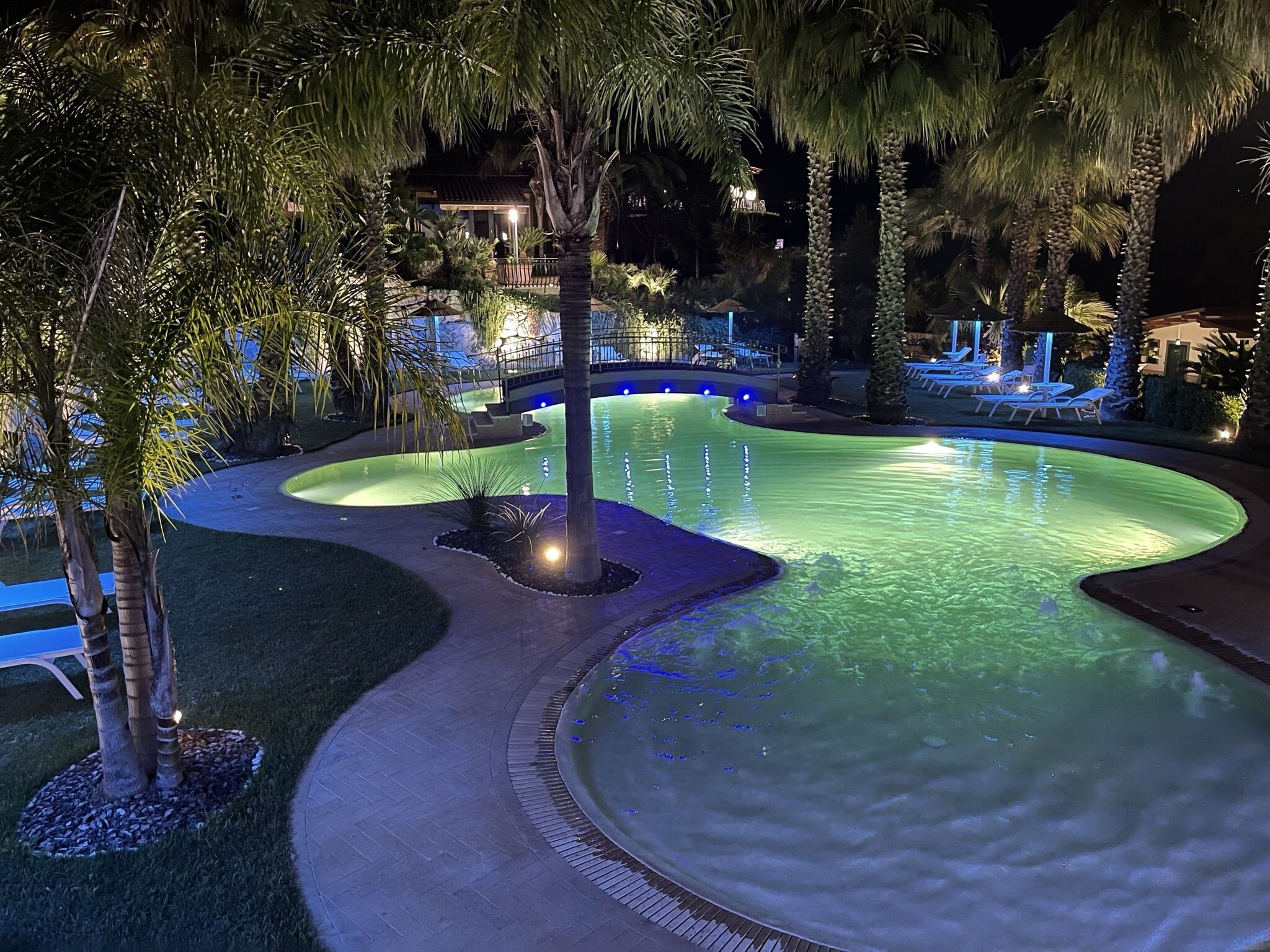Ein schwimmender Pool bei Nacht mit gebogenem Design, umgeben von Palmen und Liegestühlen.