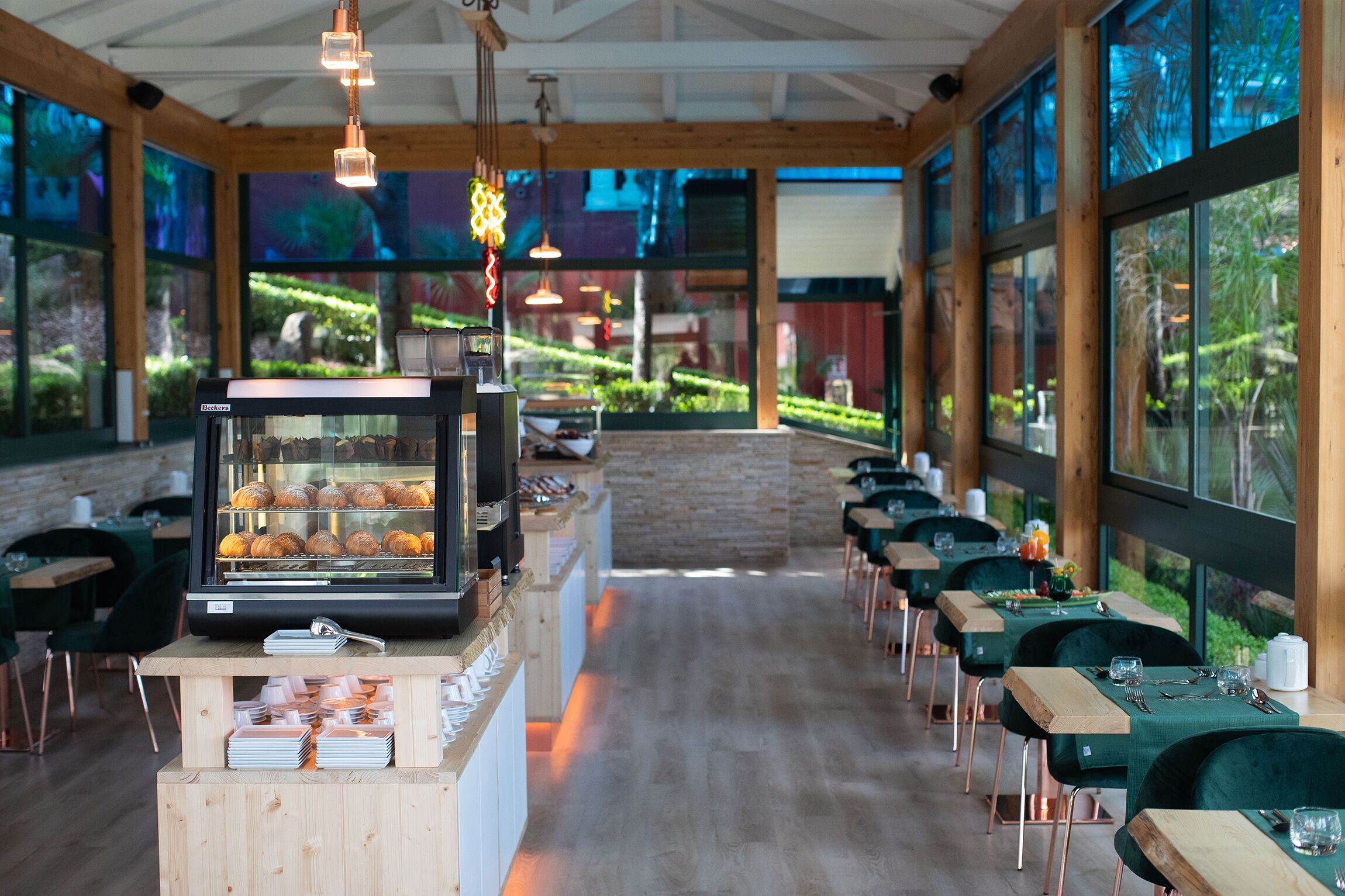 Ein Restaurant mit einer langen Theke, Holztischen und -stühlen. Eine Vitrine zeigt Gebäck, und eine Kaffeemaschine steht bereit.