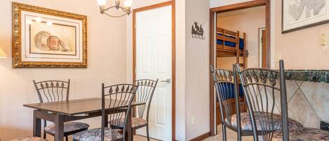 Un comedor con mesa y sillas, una araña de techo y una puerta que da a un dormitorio con literas.