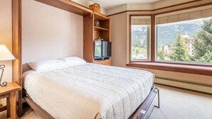 Een slaapkamer met een bed, een televisie, een houten plank met decoratieve objecten, en een raam met uitzicht op de bergen.