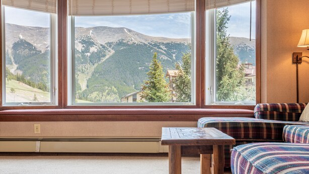 Een kamer met een groot raam met uitzicht op een bergachtig landschap, een houten tafel en een zithoek met ruitpatroon.
