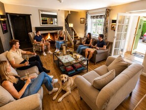 Eine Gruppe von Personen sitzt auf Sofas in einem Wohnzimmer mit Kamin, einem Hund und einem Couchtisch mit Gesellschaftsspielen.