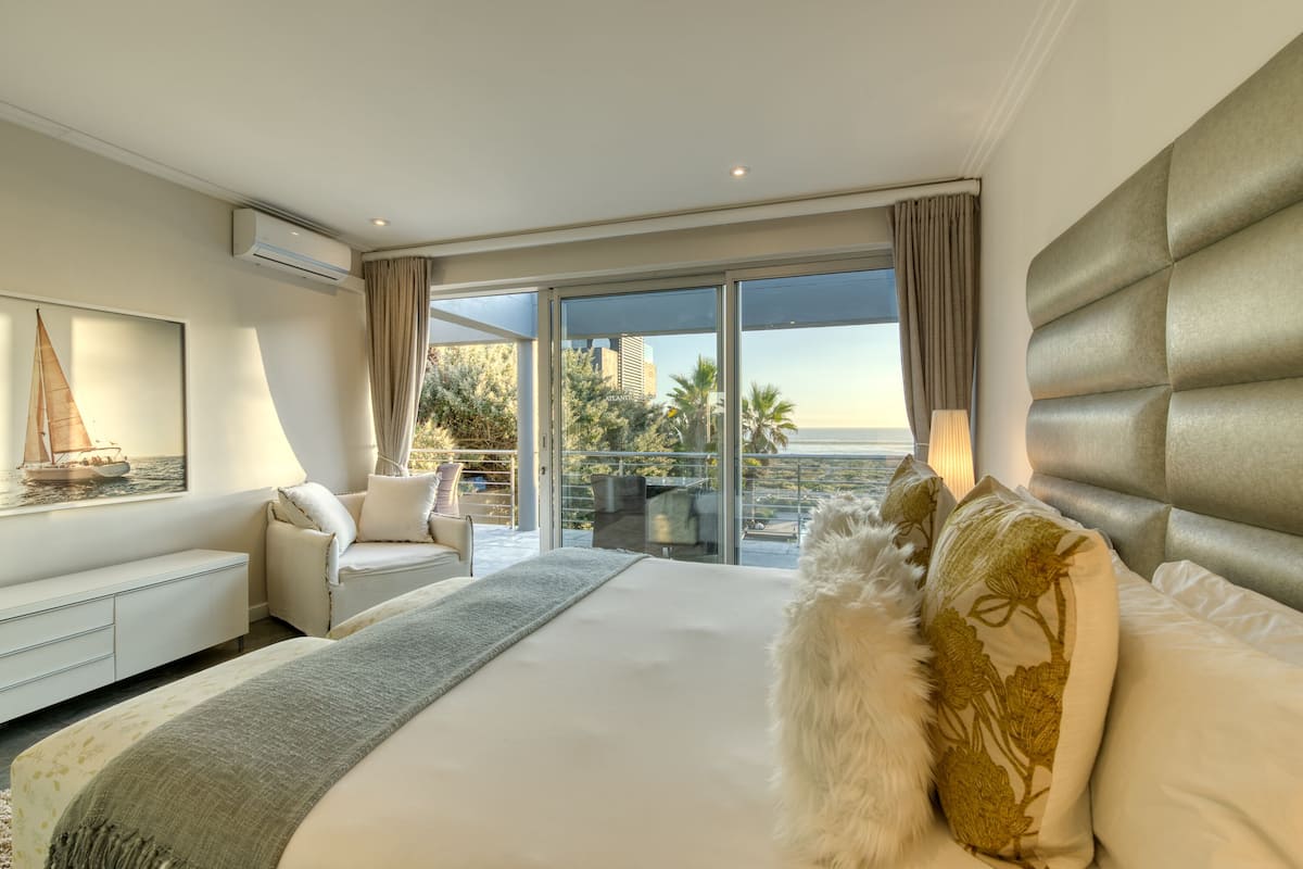 A modern hotel room with a large bed, a sofa, and a balcony with a view of palm trees and buildings.