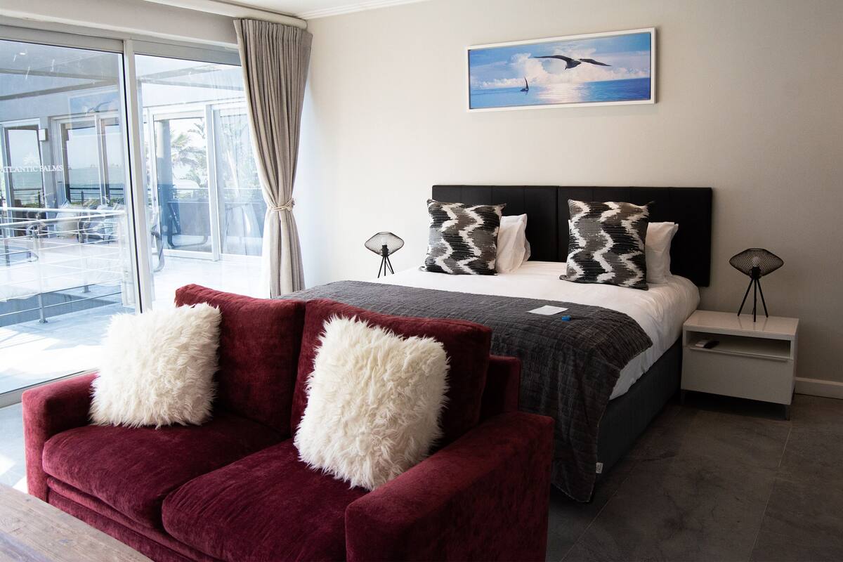 A modern hotel room with a large bed, two nightstands, a red sofa with white cushions, and a view of the ocean through large windows.