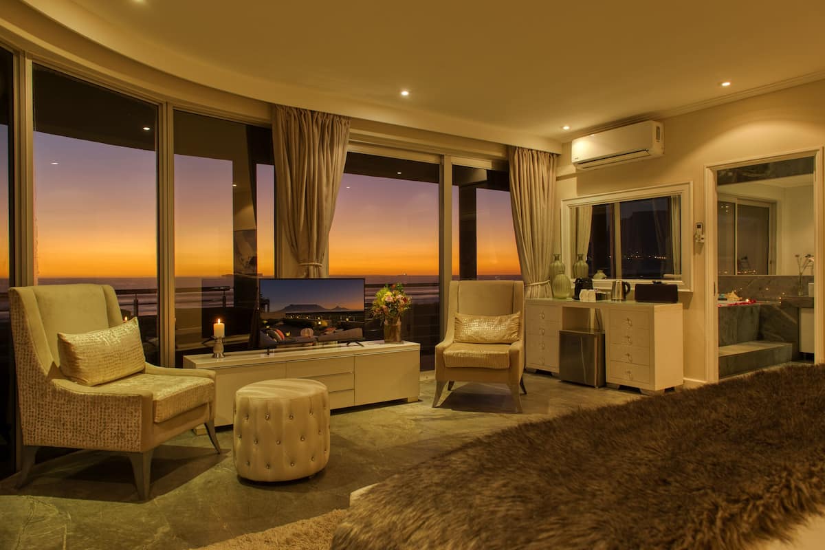 A spacious living room with a large TV, comfortable seating, a small kitchenette, and a view of the sunset over the water.