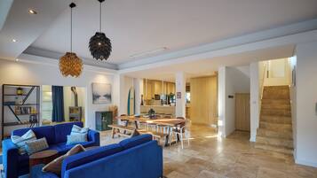 A spacious living area with a blue sofa set, a wooden dining table, and a kitchen in the background.