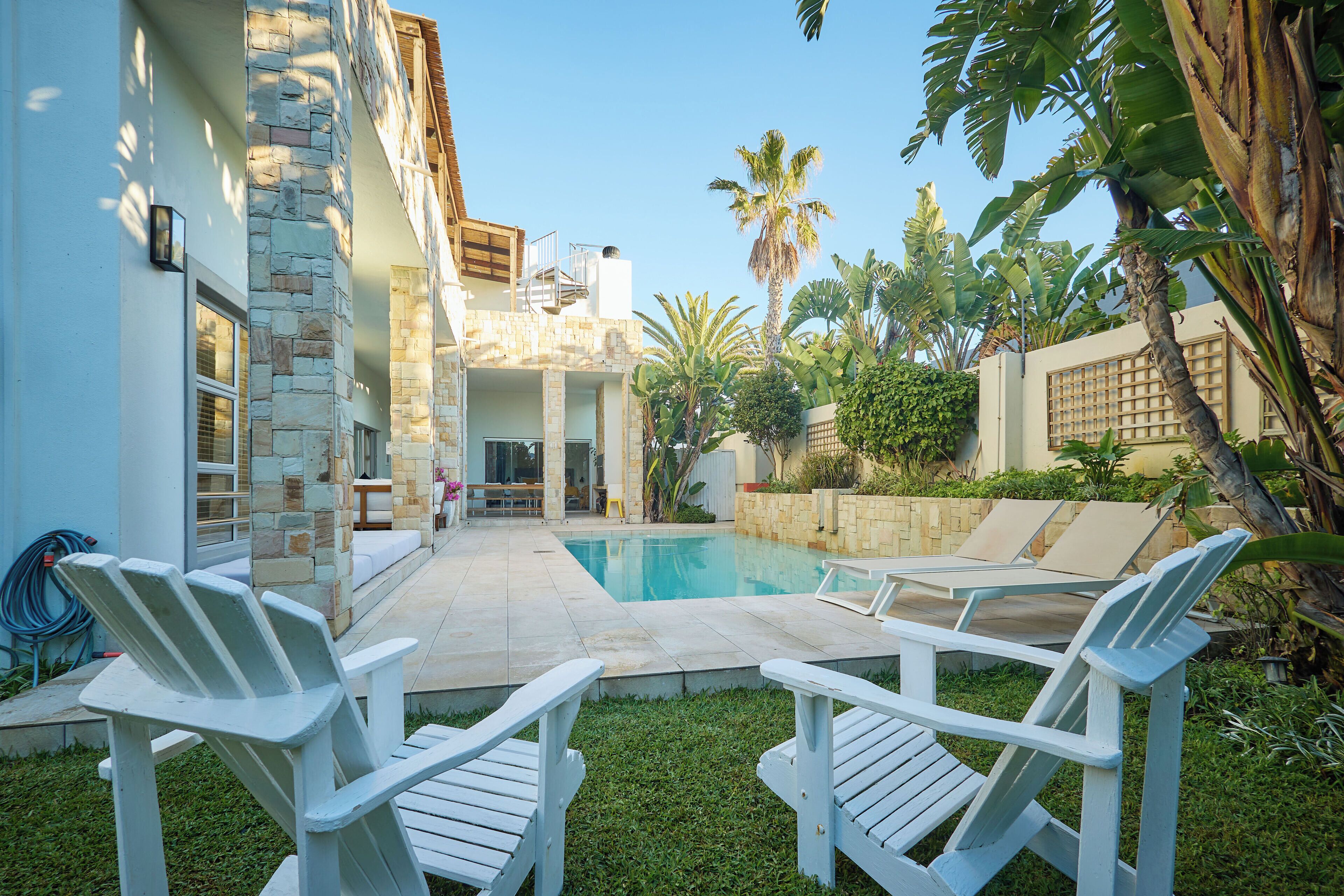 A pool area with two white Adirondack chairs, a stone building, and tropical plants.
