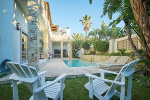 A pool area with two white Adirondack chairs, a stone building, and tropical plants. - Atlantic Palms (Cape Town)