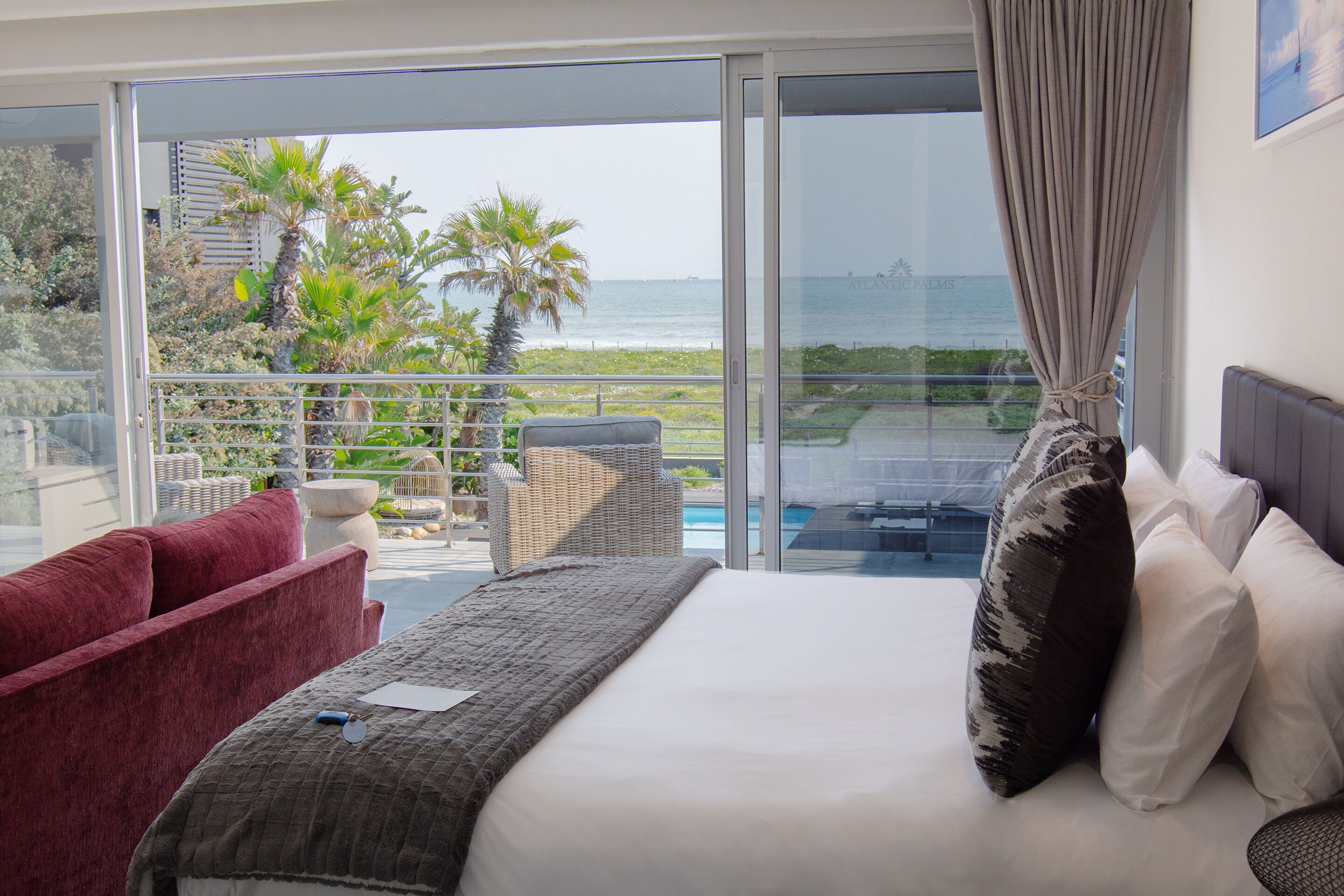 Habitación de hotel con cama, sofá rojo, sillón de mimbre y vistas al océano y vegetación.