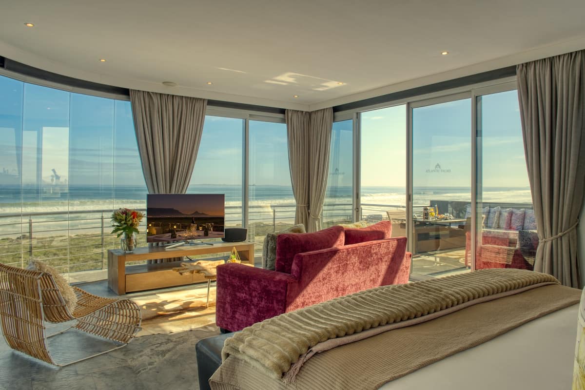A modern hotel room with a large balcony, a flat-screen TV, a sofa, and a dining area with a view of the beach.