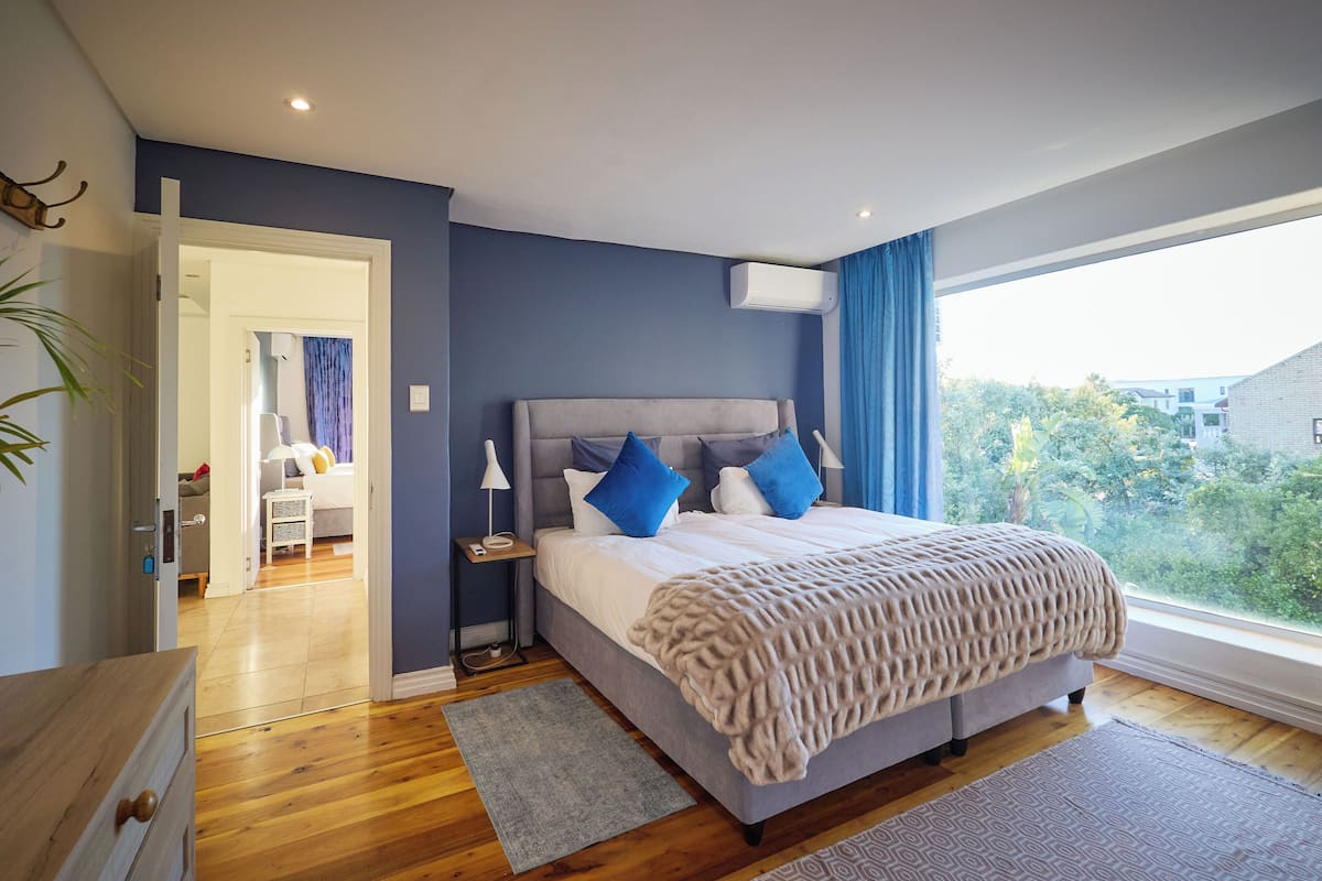 A bedroom with a bed, bedside tables, a nightstand, a window with curtains, and a view of greenery outside.