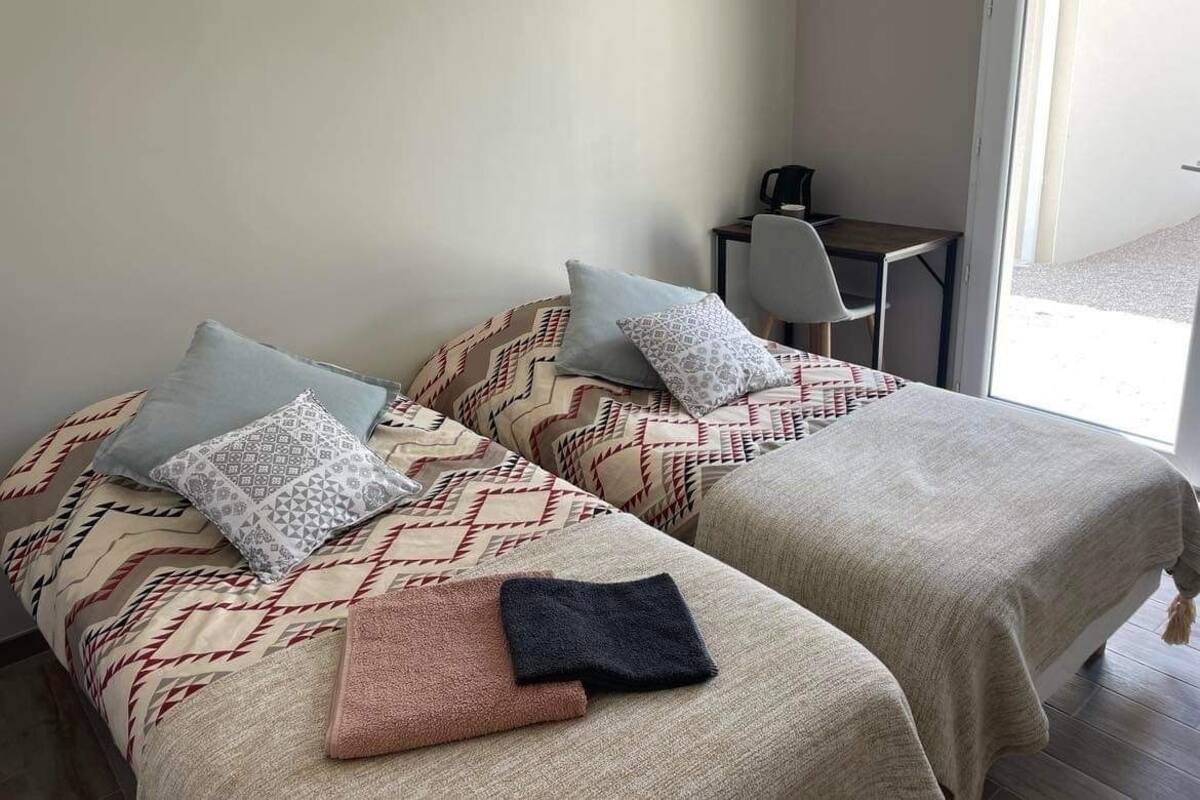 Two beds with patterned bedding and pillows, a small table with a chair, and a window providing natural light.