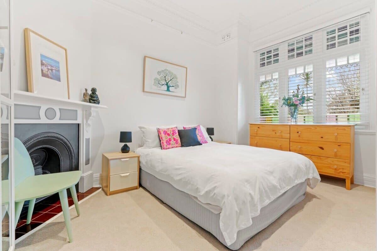 A bedroom with a bed, dresser, nightstand, chair, fireplace, and window with blinds.