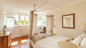 A bedroom with a four-poster bed, a wooden dresser, a mirror, and a view of greenery outside.