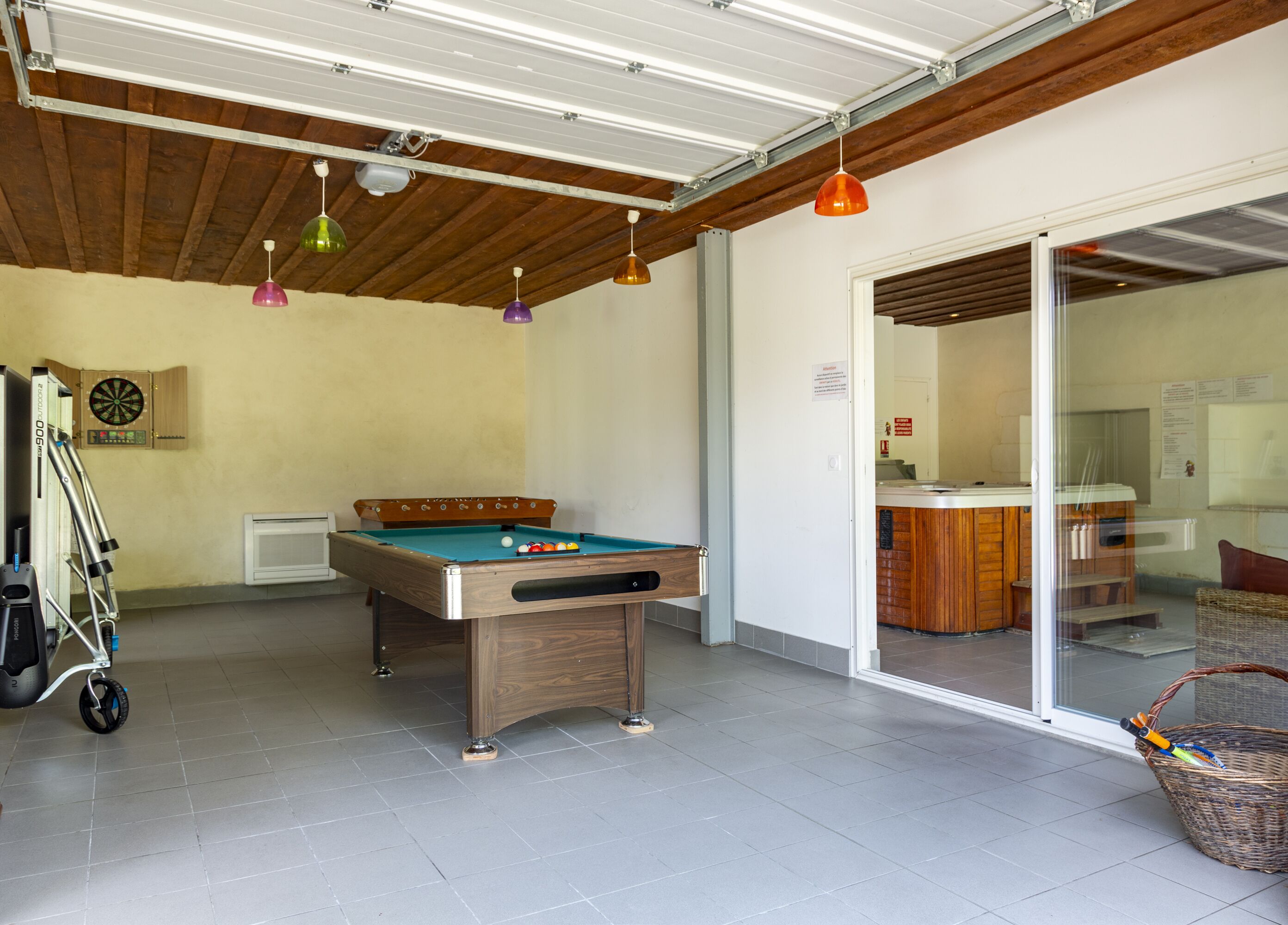A pool table with a dartboard and a foosball table in a room with a sliding glass door leading to a bar area.