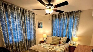 A hotel room with a bed, two bedside tables, a ceiling fan, patterned curtains, a rug, and a tiled floor.
