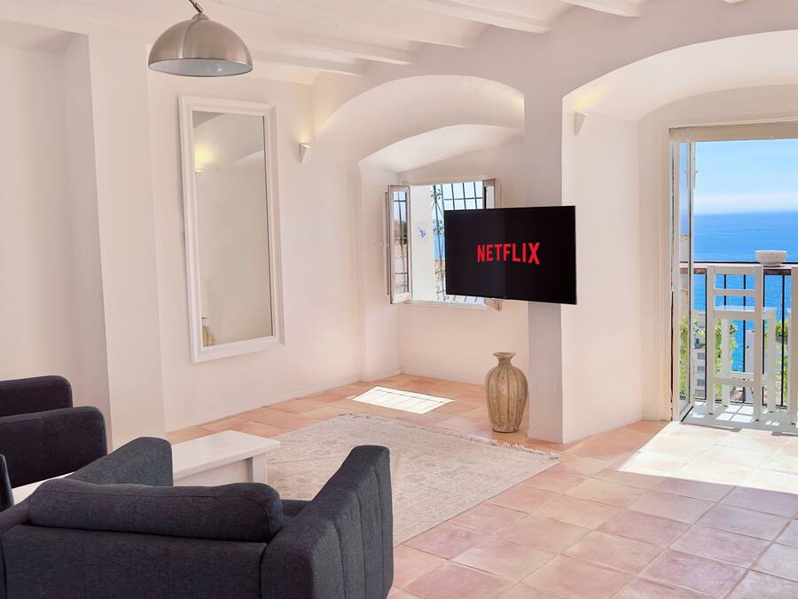 A living room with a flat-screen TV mounted on the wall, a sofa, a coffee table, and a view of the sea through an open balcony door.
