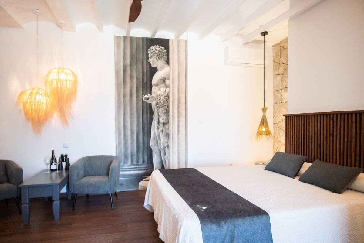 A modern hotel room with a large bed, a seating area with a table and chairs, a statue, and a stone wall.