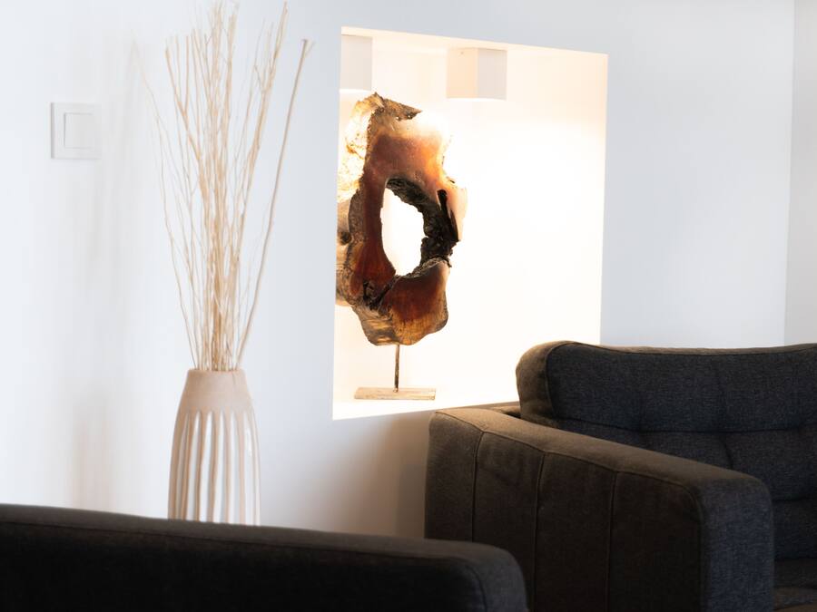 Una sala de estar moderna con un sofá gris oscuro, un florero blanco con plantas secas y una escultura abstracta única sobre un pedestal blanco.