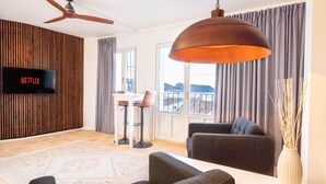 A modern living room with a flat-screen TV, a ceiling fan, a large pendant light, a sofa, a coffee table, and a view of the outdoors through large windows. - Porta Nova Suites Altea - Adults Only (Altea)