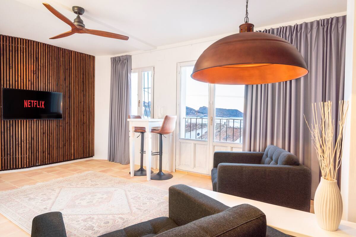 A modern living room with a flat-screen TV, a ceiling fan, a large pendant light, a sofa, a coffee table, and a view of the outdoors through large windows.