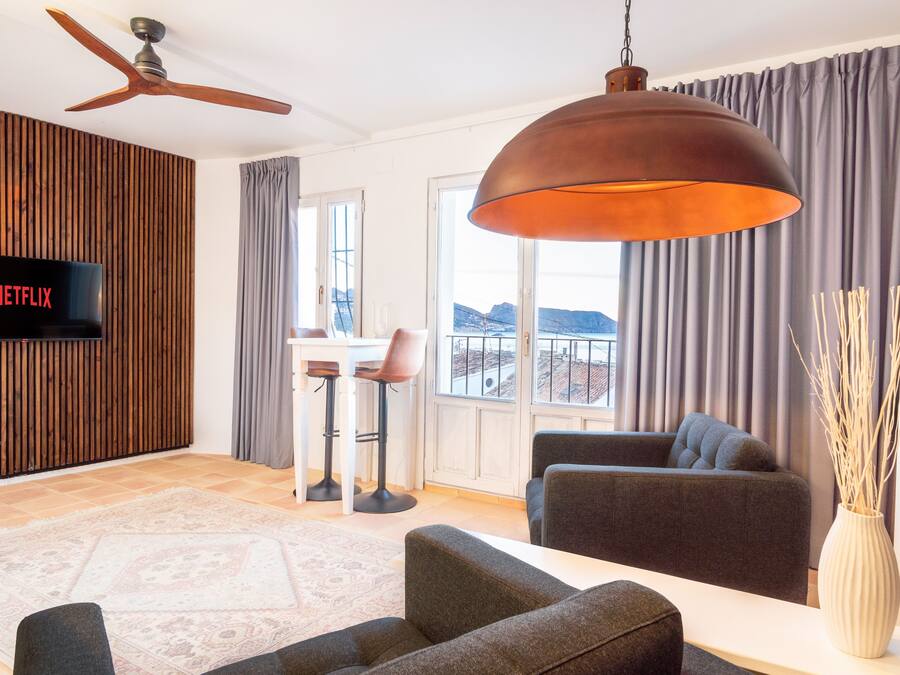 Una sala de estar moderna con una televisión de pantalla plana, un ventilador de techo, una lámpara colgante grande, un sofá, una mesa de centro y vistas al exterior a través de amplios ventanales.