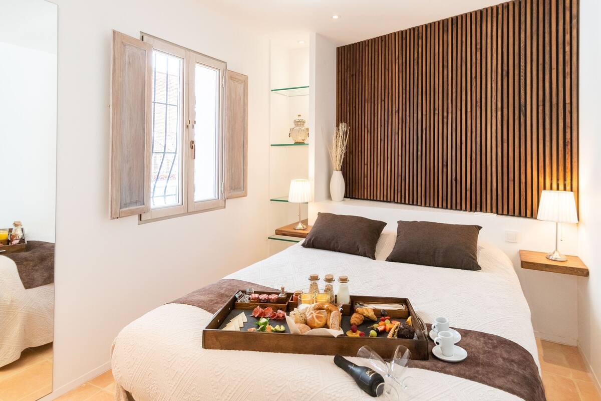 A hotel room with a large bed, a tray of breakfast items, a wooden wall panel, and a bedside table with a lamp.
