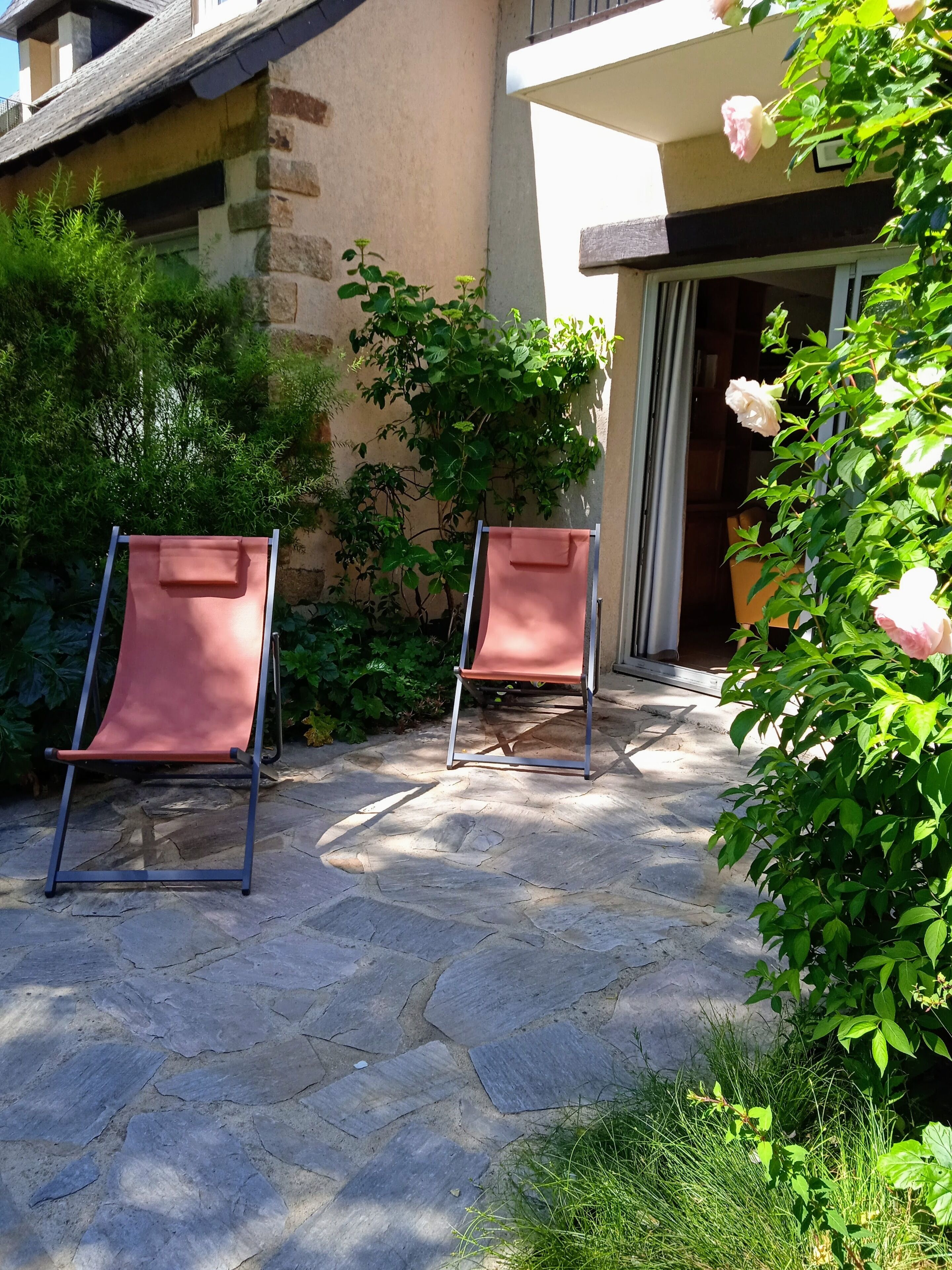Two outdoor chairs on a stone patio with a building in the background and greenery around.