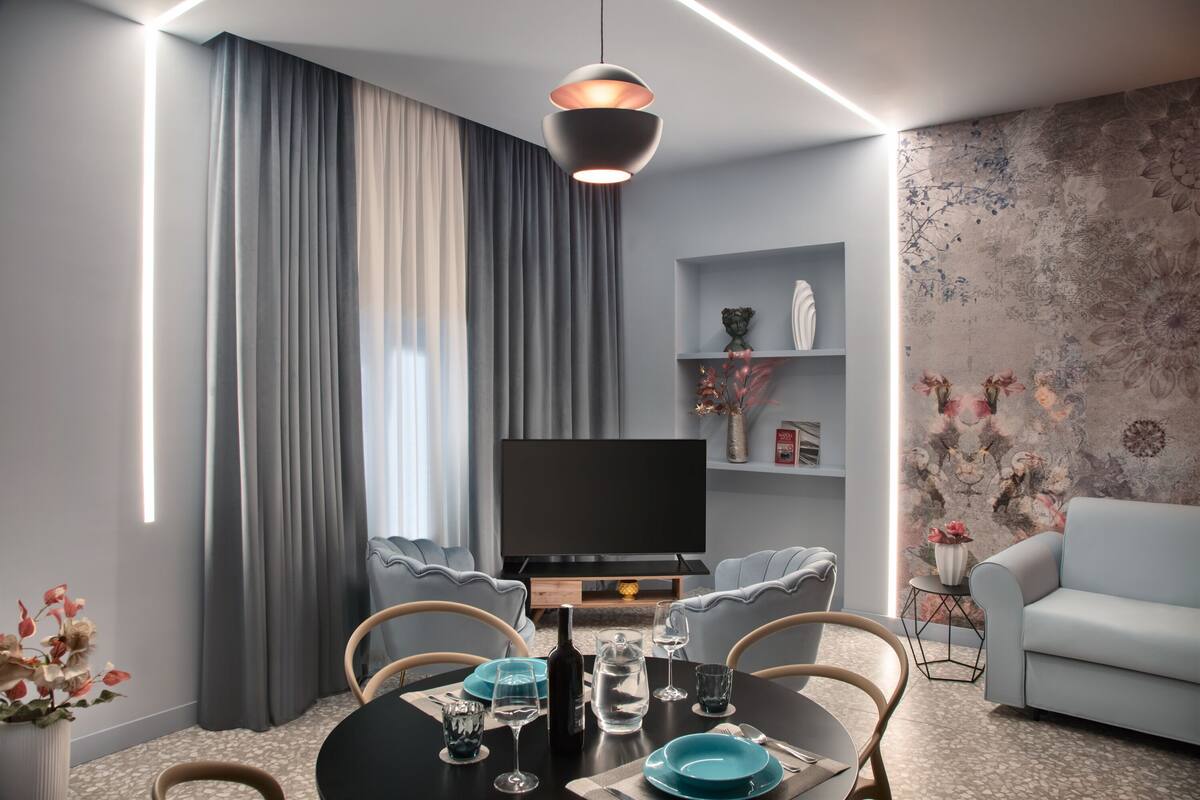 Ett modernt vardagsrum med en matplats, grå gardiner, en platt-TV och en vägg med ett blommönster.
